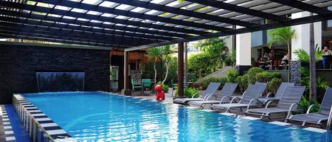 Piscina com pergolado, cadeiras de descanso e uma cascata.