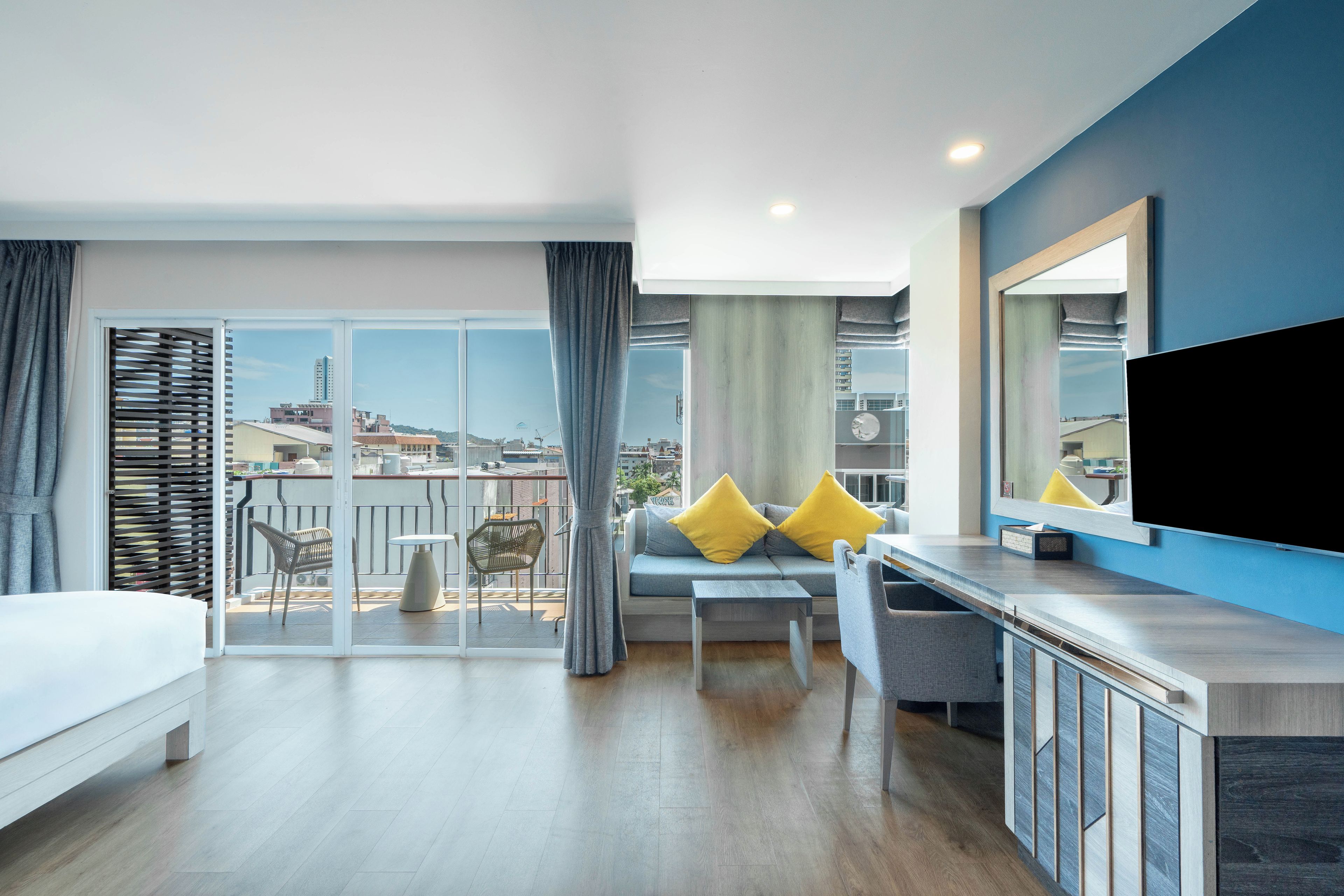 A modern hotel room with a balcony, a bed, a seating area with cushions, and a television.