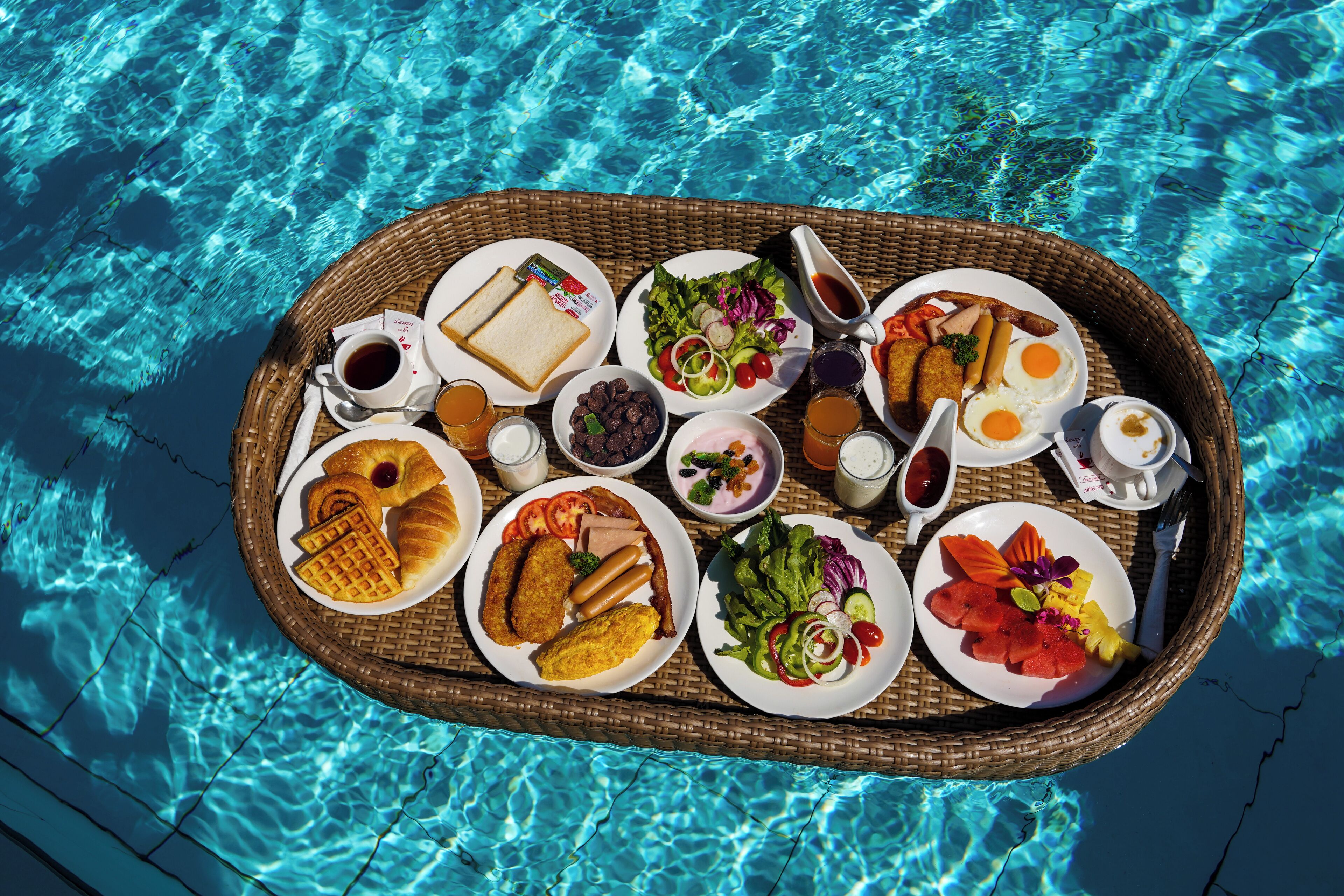 Una bandeja de mimbre con diversos productos para el desayuno, como pan, huevos, salchichas y verduras frescas, sobre una mesa junto a la piscina.