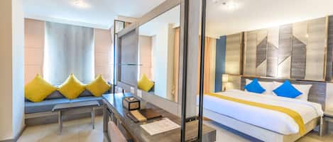 A modern hotel room with a large bed, a desk with a computer, and a seating area with cushions.