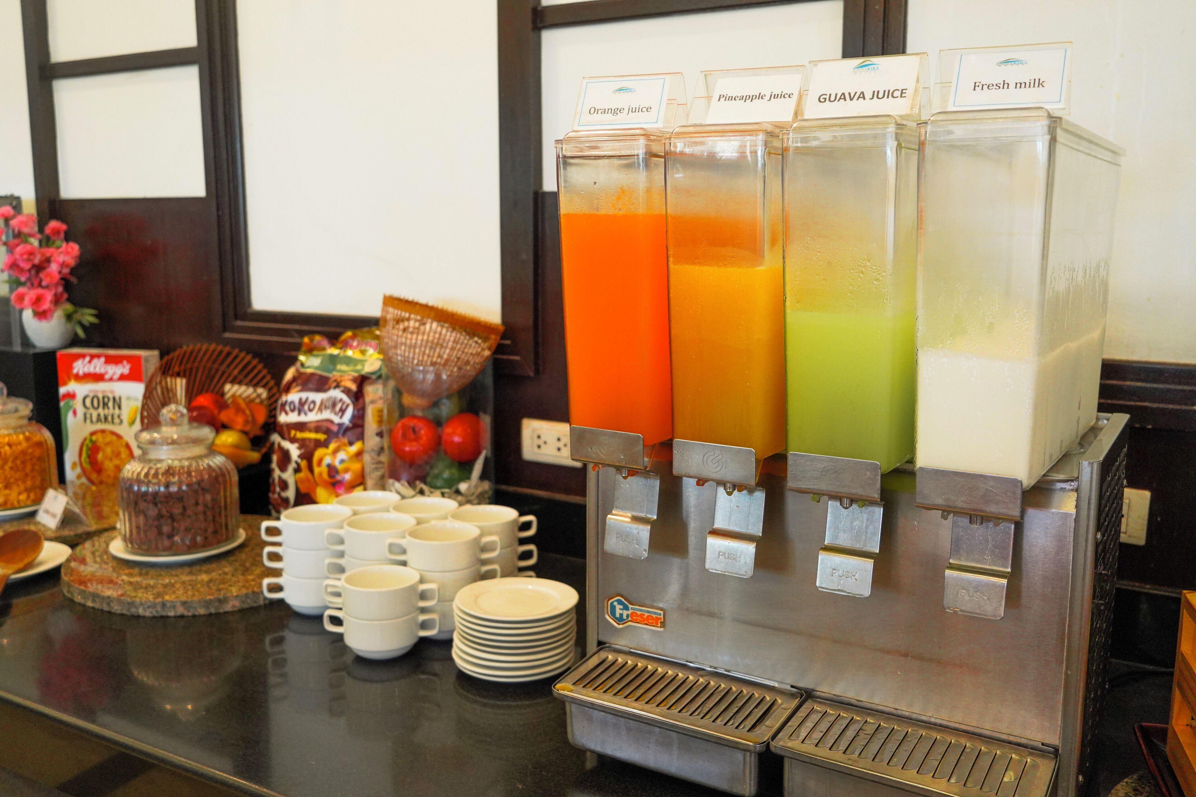 A breakfast bar with juice dispensers, cereal, and coffee mugs.