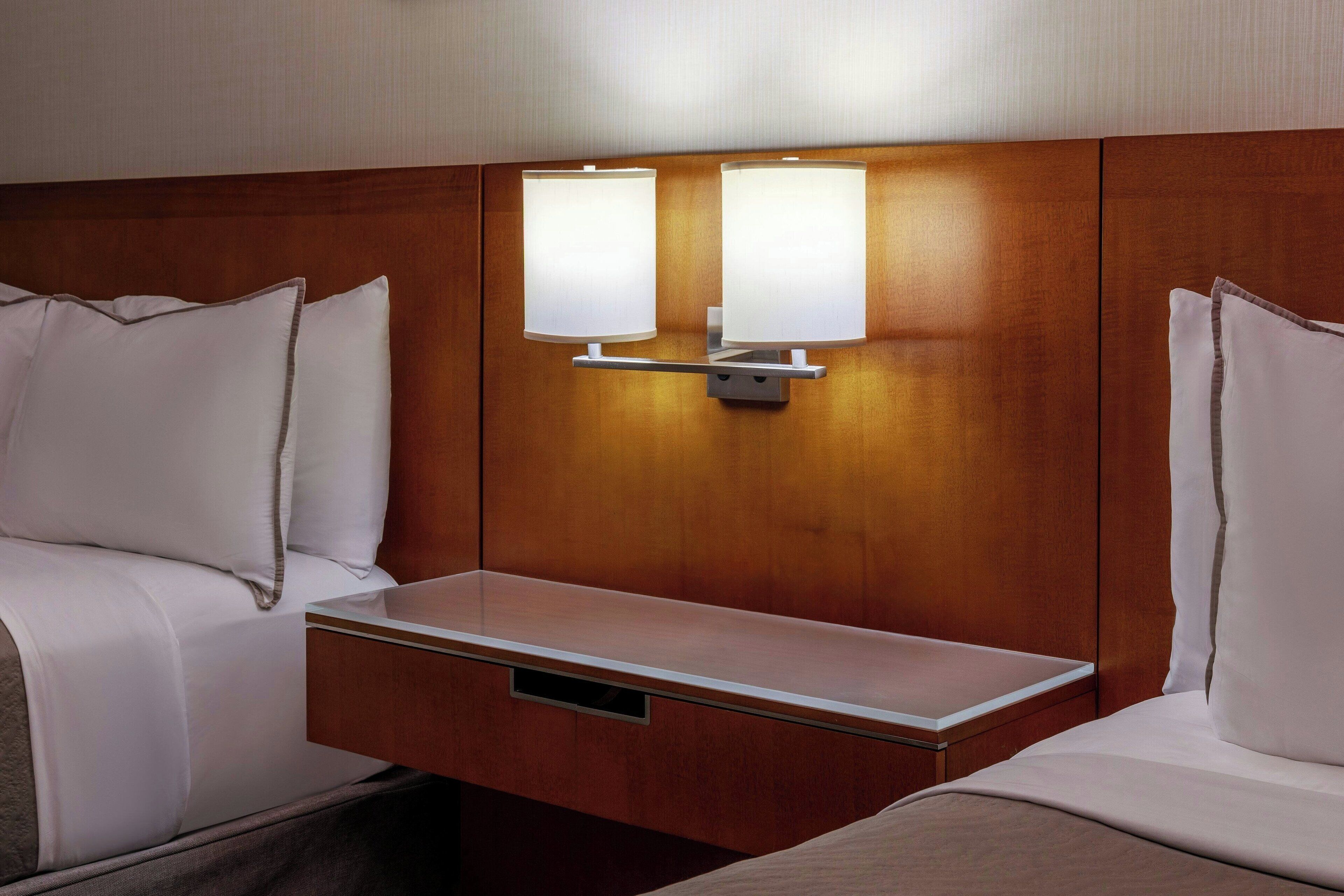 Habitación de hotel con cabecera de madera, dos mesitas de noche y dos lámparas de pared.