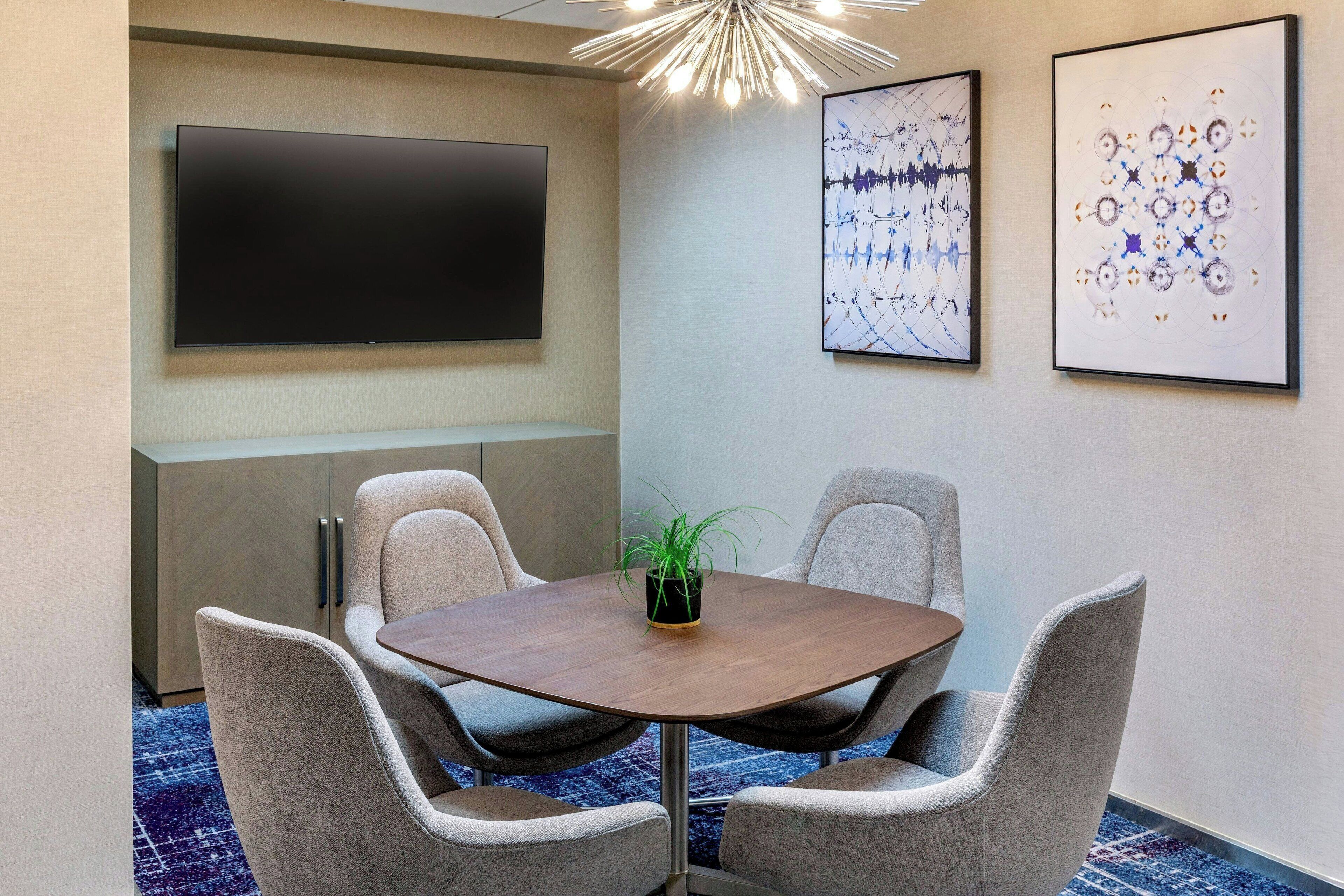 A conference room with a round table, four chairs, a potted plant, a flat-screen TV, and abstract wall art.