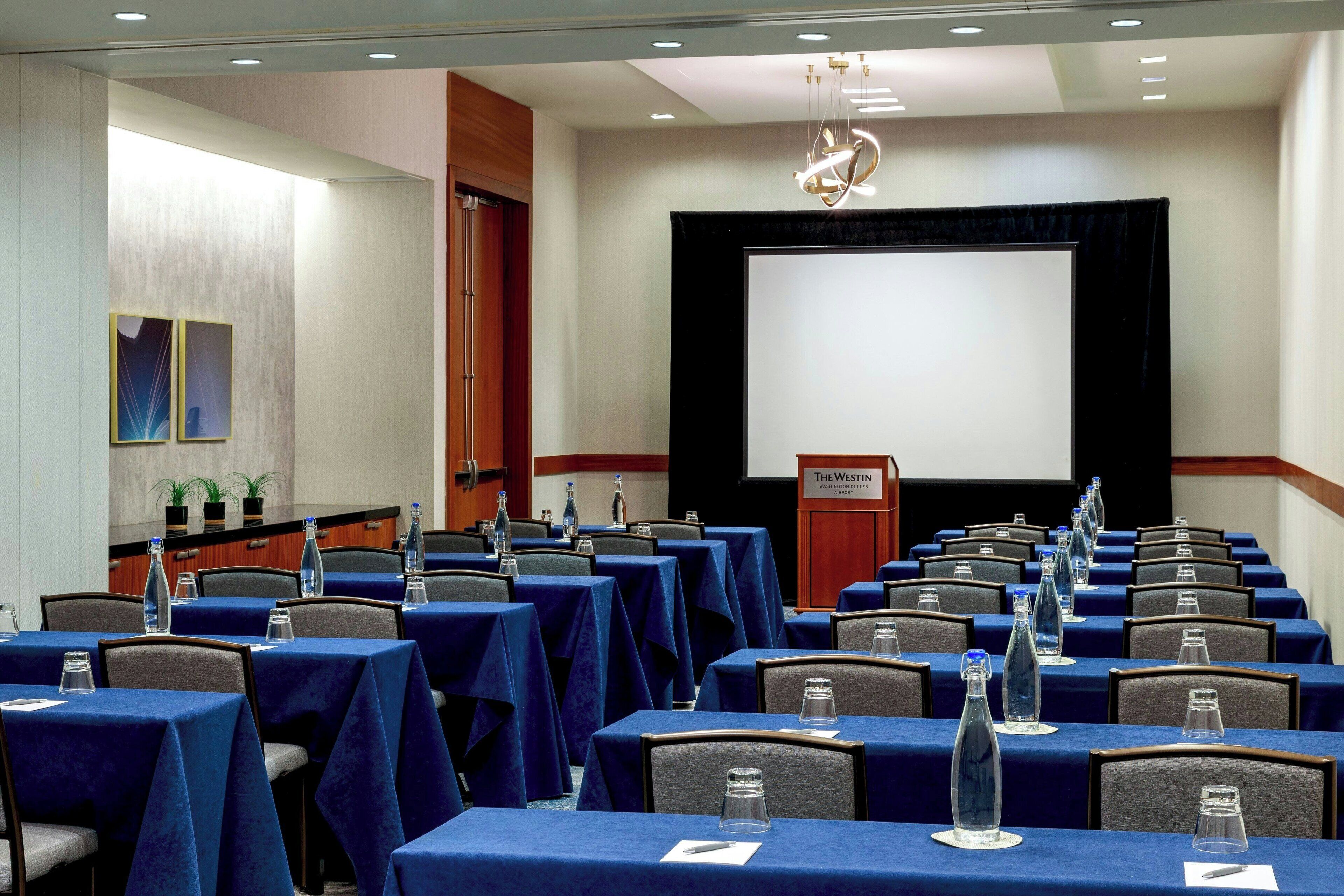 Una sala conferenze con file di tavoli, sedie e un grande schermo di fronte.