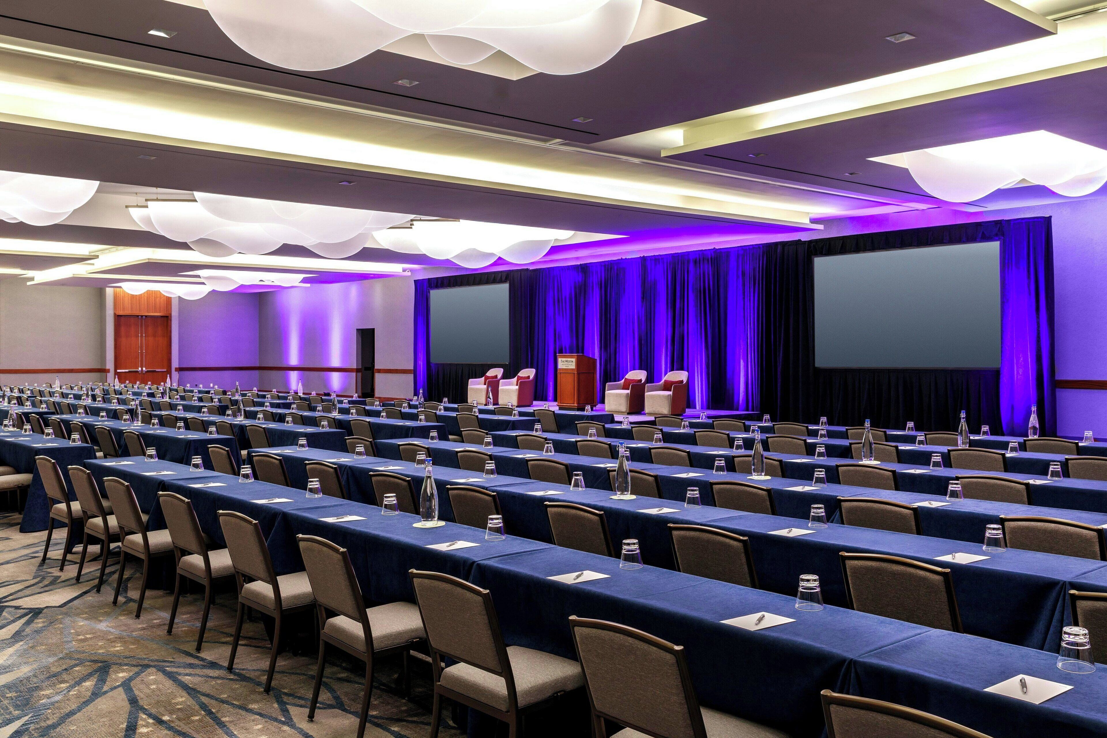 A conference room with rows of tables and chairs, a large screen, and a podium.