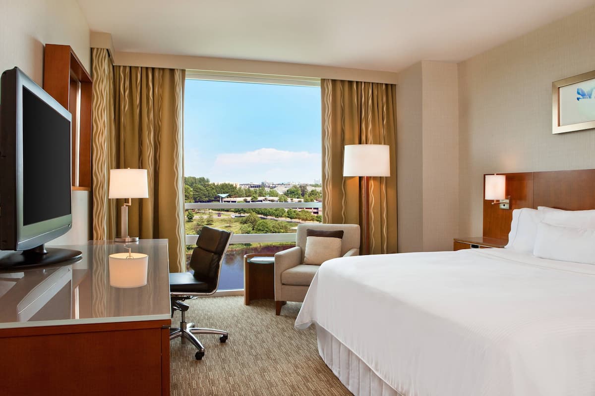 A hotel room with a large bed, a desk with a TV, a chair, and a view of a cityscape through the window.