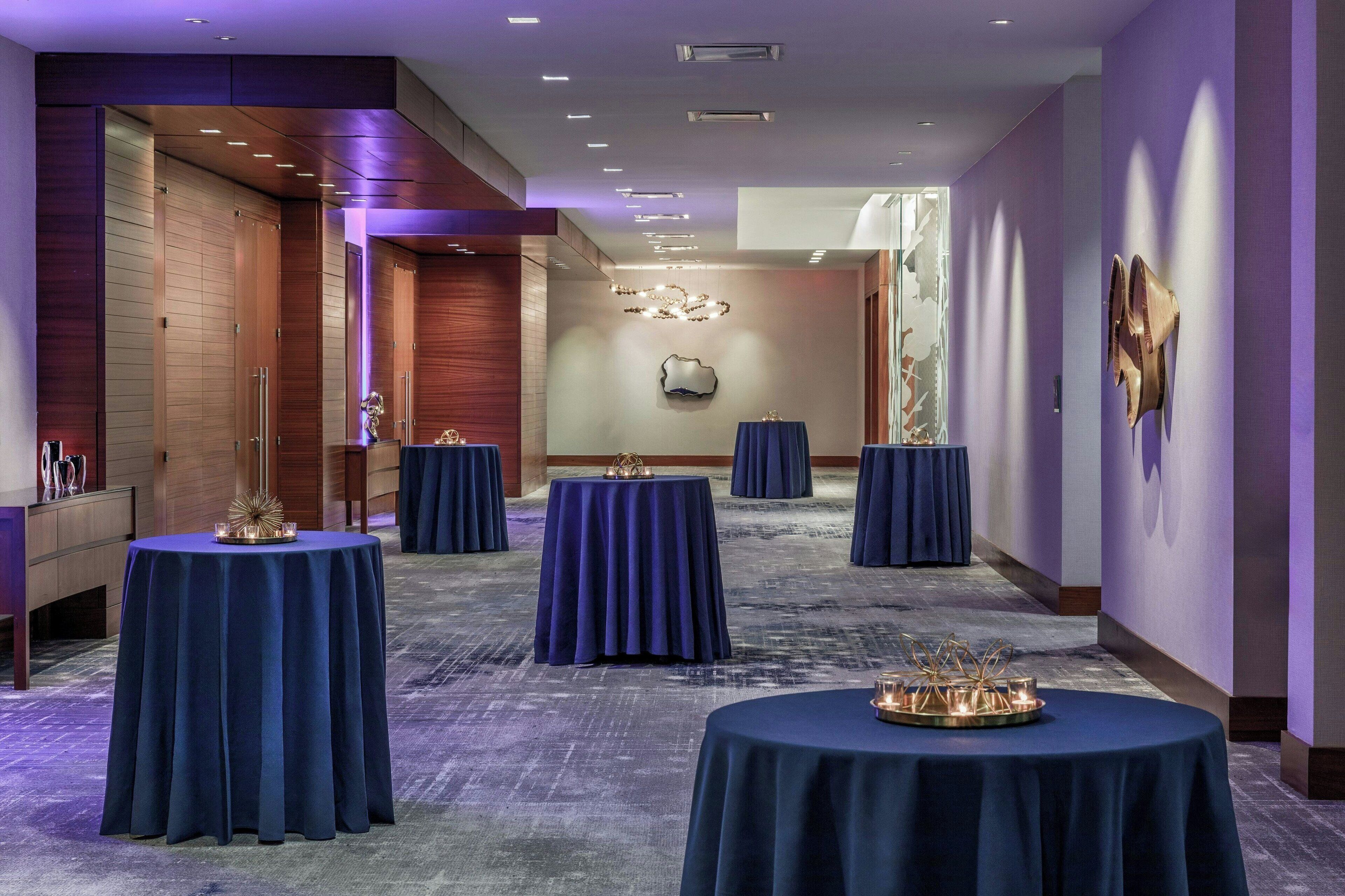 A hotel lobby with multiple round tables covered with blue tablecloths, a chandelier, and a large mirror on the wall.