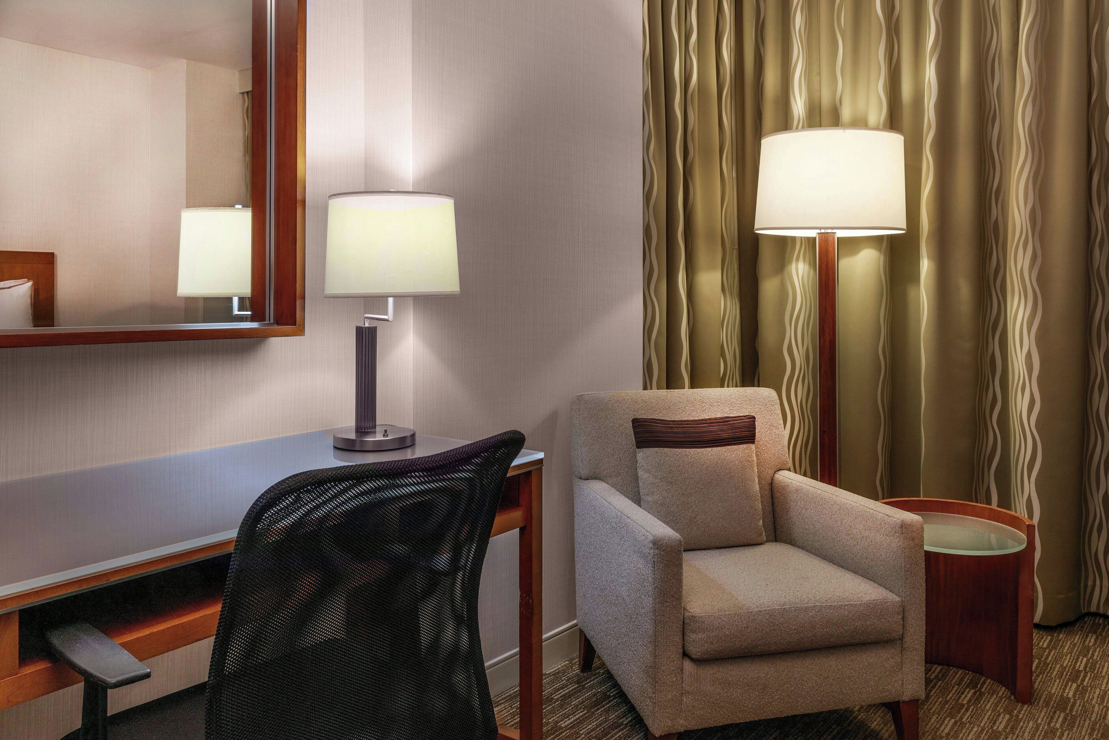 A hotel room with a desk, chair, lamp, and a large mirror.