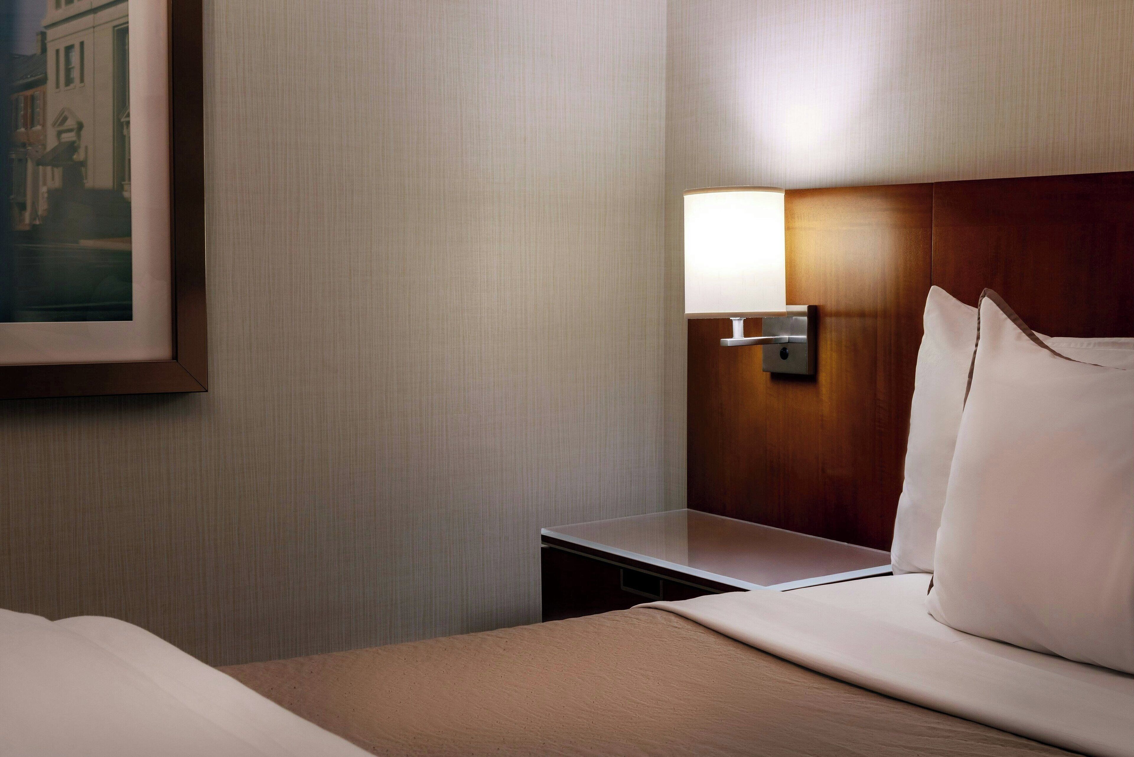 A hotel room with a neatly made bed, a bedside table, and a wall-mounted lamp.