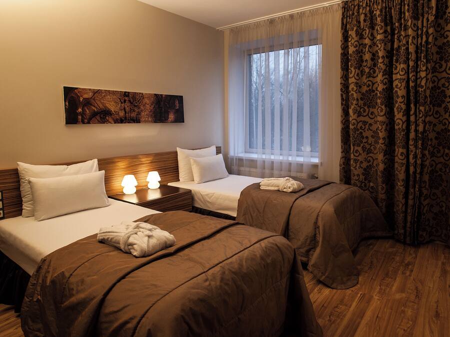 A hotel room with two beds, a nightstand with a lamp, a painting on the wall, and a window with curtains.
