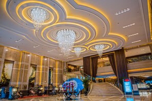 Un amplio vestíbulo con una imponente escalera, múltiples candelabros y una zona de recepción con asientos y un panel informativo digital.