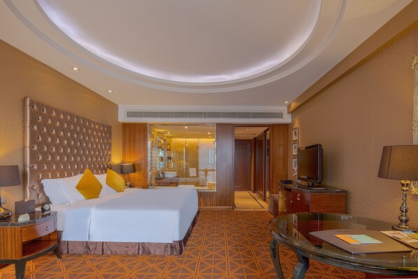Une chambre d’hôtel avec un grand lit, une table en verre, un téléviseur et une salle de bain visible à travers une porte ouverte.