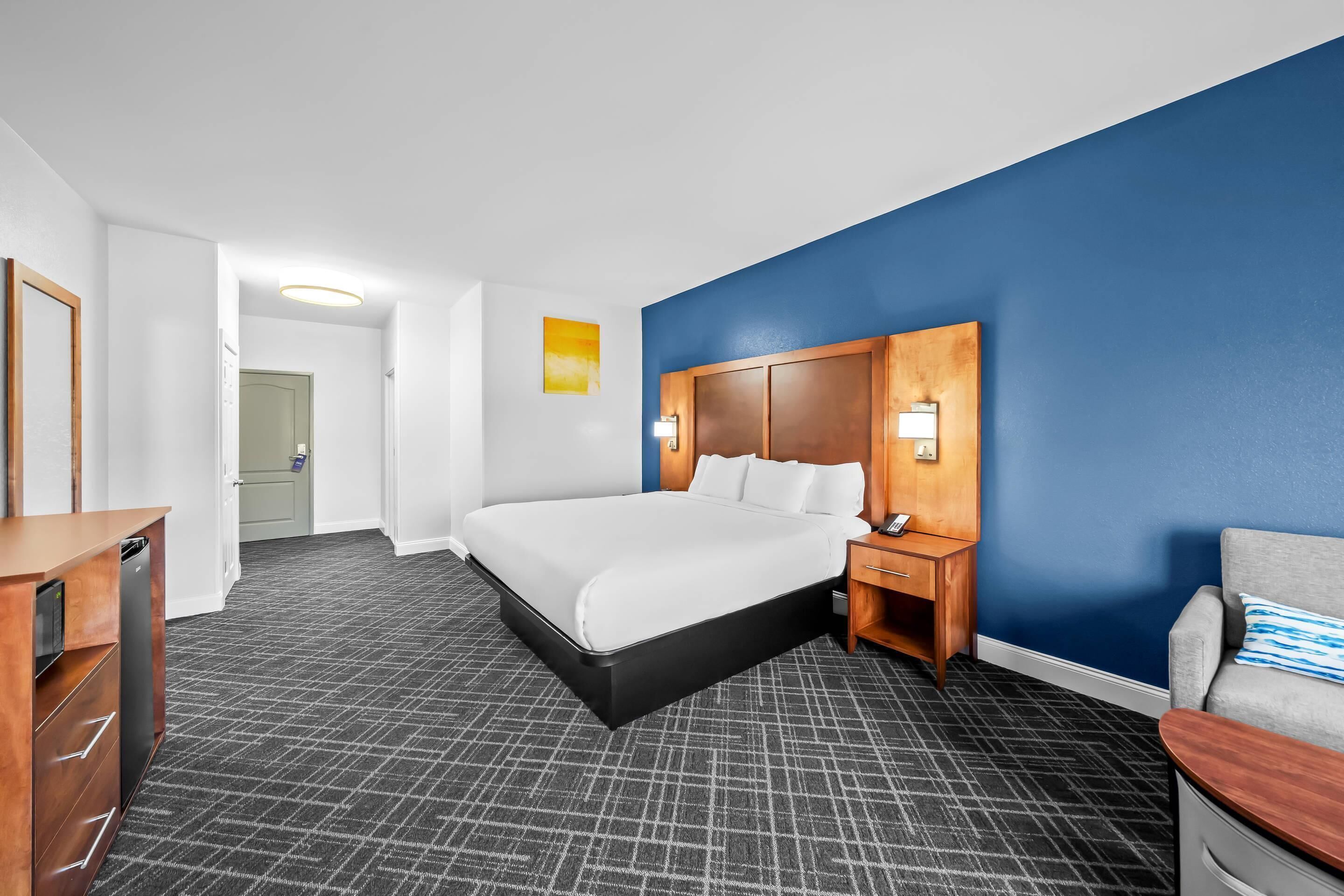 Une chambre d’hôtel avec un grand lit, une table de chevet en bois, un bureau et une chaise. Une porte donne accès à une autre pièce.