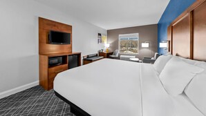 Hotellihuone, jossa on suuri sänky, seinään kiinnitetty litteänäyttöinen televisio, kirjoituspöytä tuolilla, pieni pöytä lampun kanssa ja ikkuna, josta avautuu näkymä rakennuksiin.