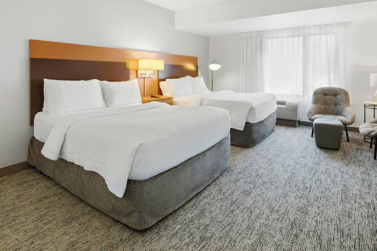 A hotel room with two beds, a chair, a lamp, and a window with curtains.