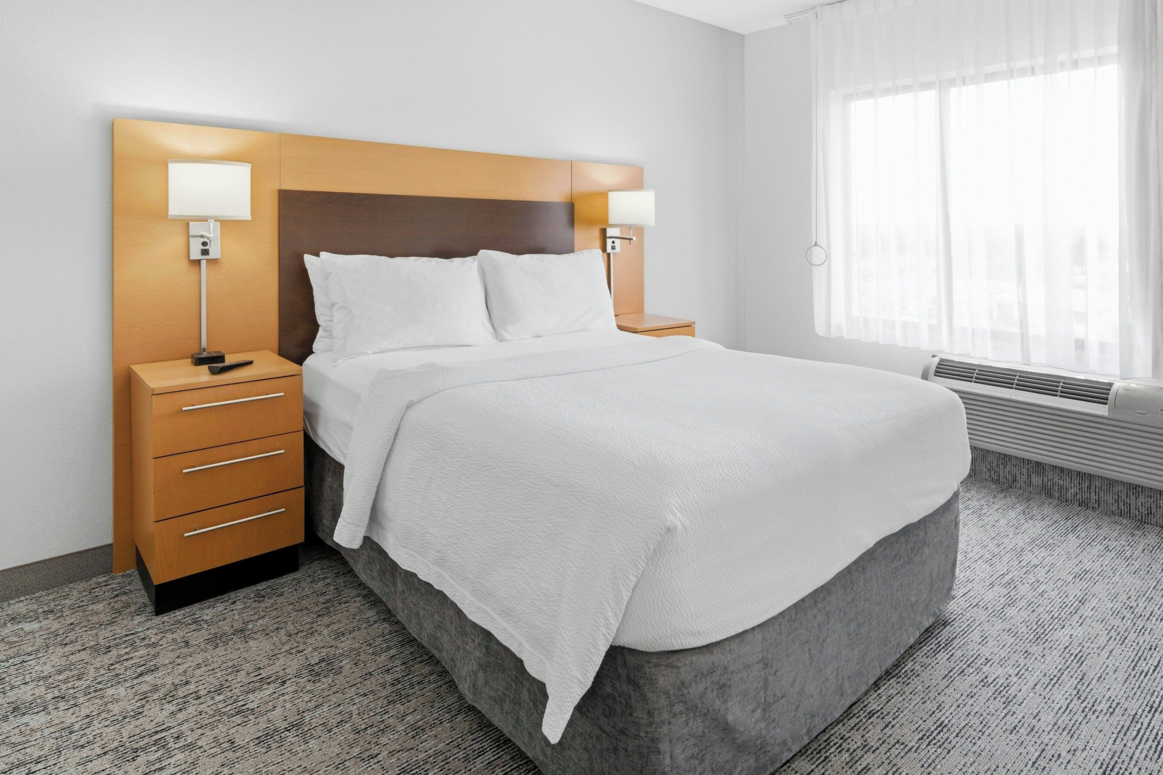 A hotel room with a large bed, two bedside tables with lamps, a wooden headboard, and a large window with sheer curtains.