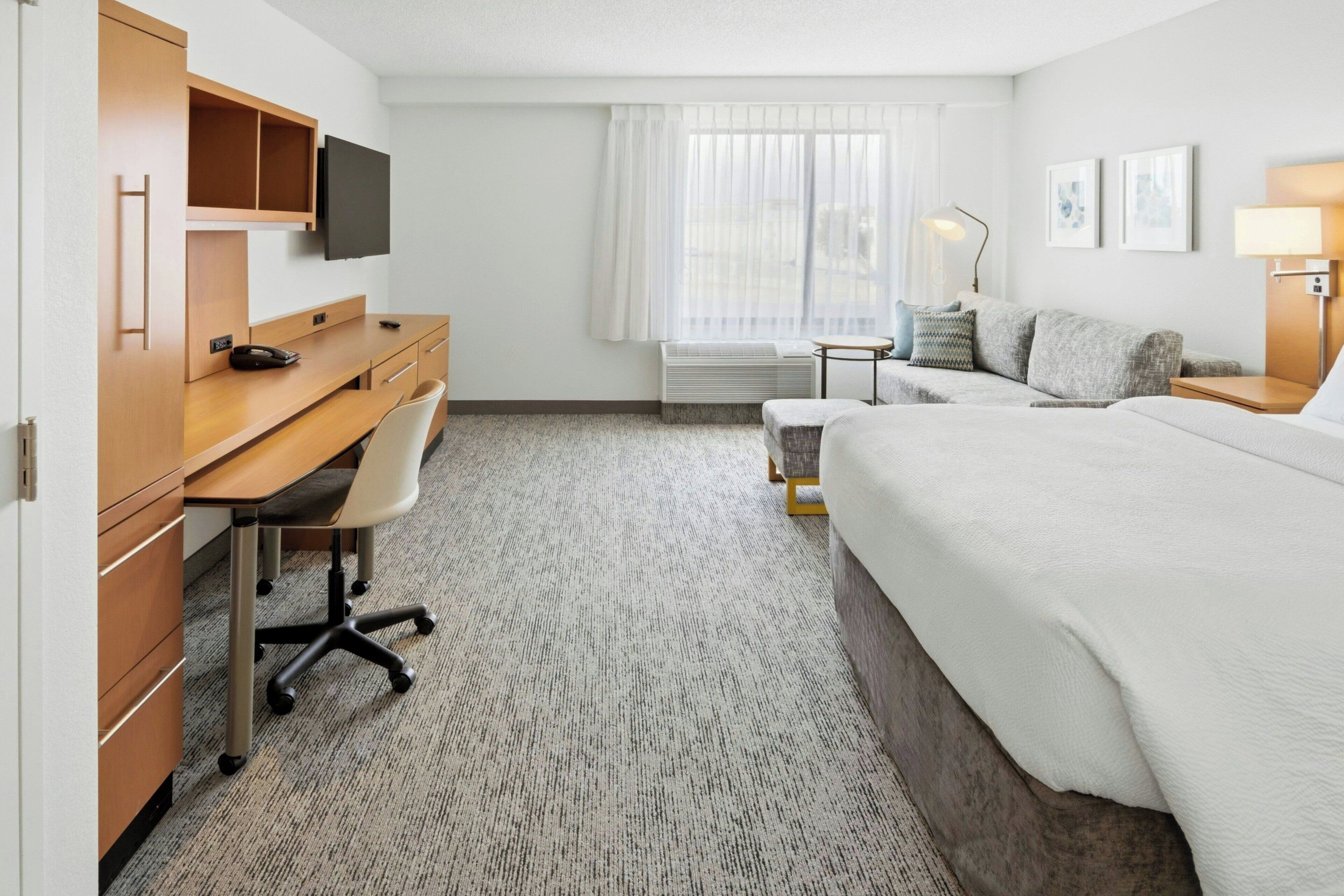 Habitación de hotel con cama, escritorio, silla, televisor, aire acondicionado y ventana con cortinas.