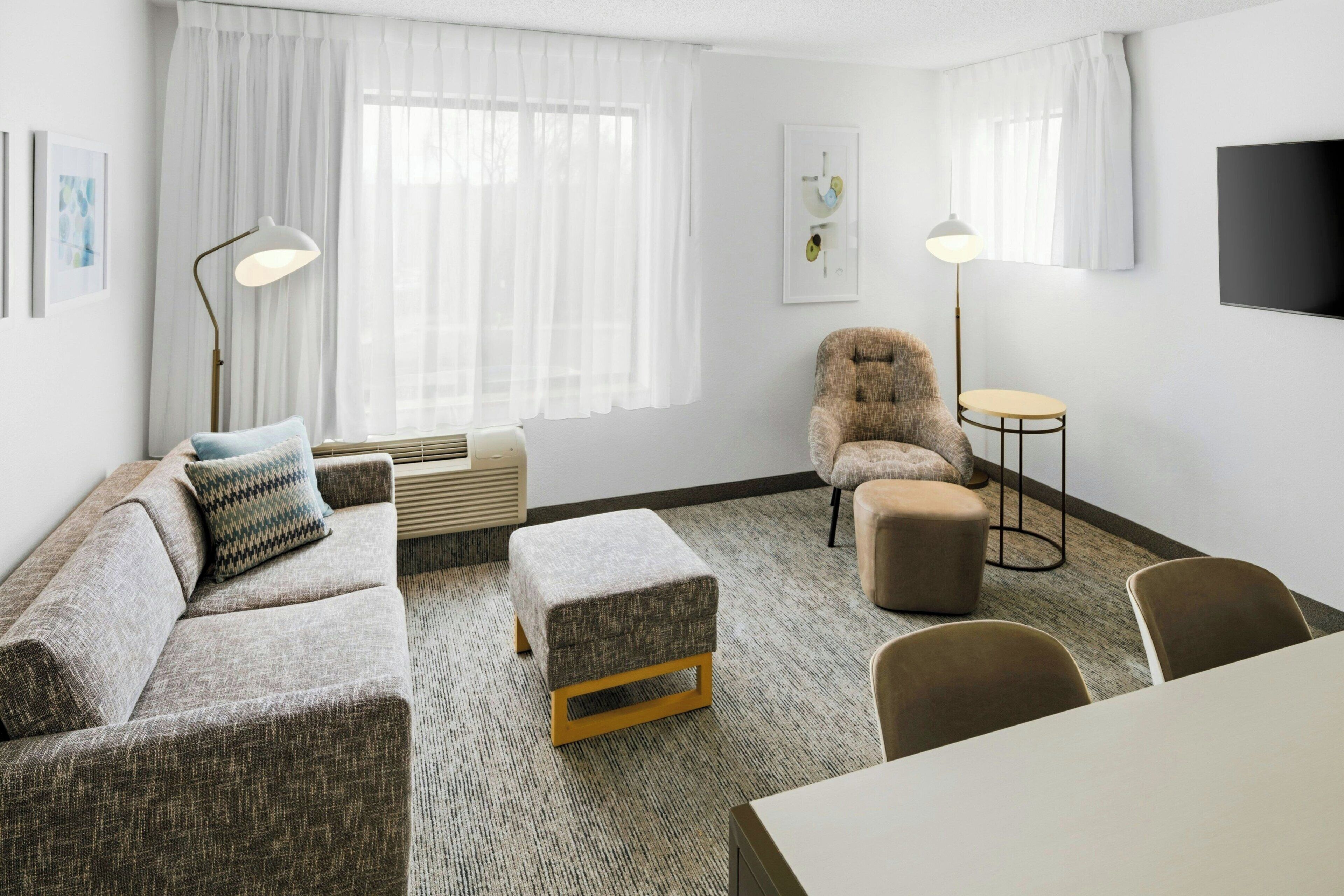 Quarto de hotel moderno com sofá, banquinho, cadeira, uma mesinha redonda e uma TV fixada na parede.