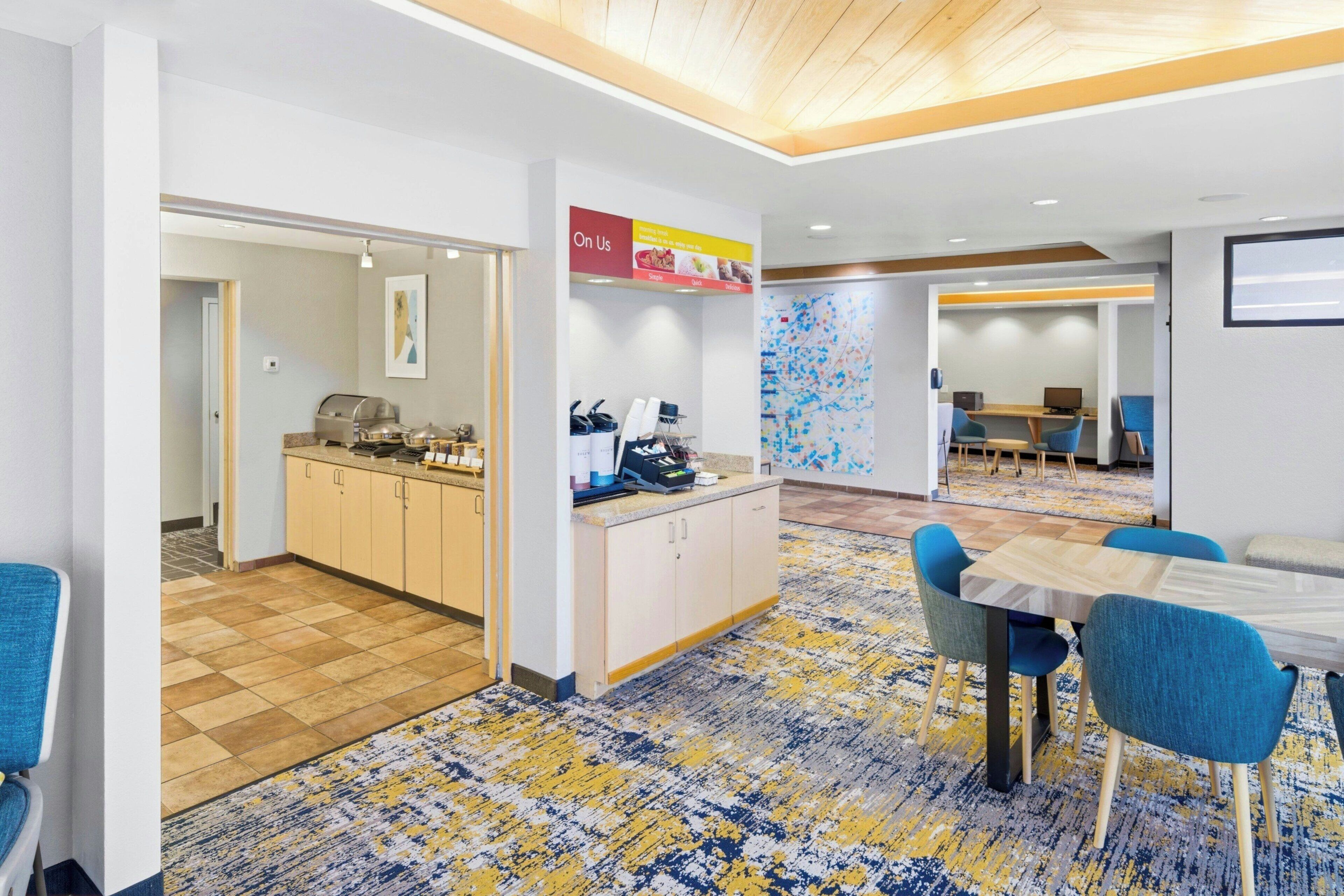 A modern hotel lobby with a kitchen area, a dining area with blue chairs, and a patterned carpet.