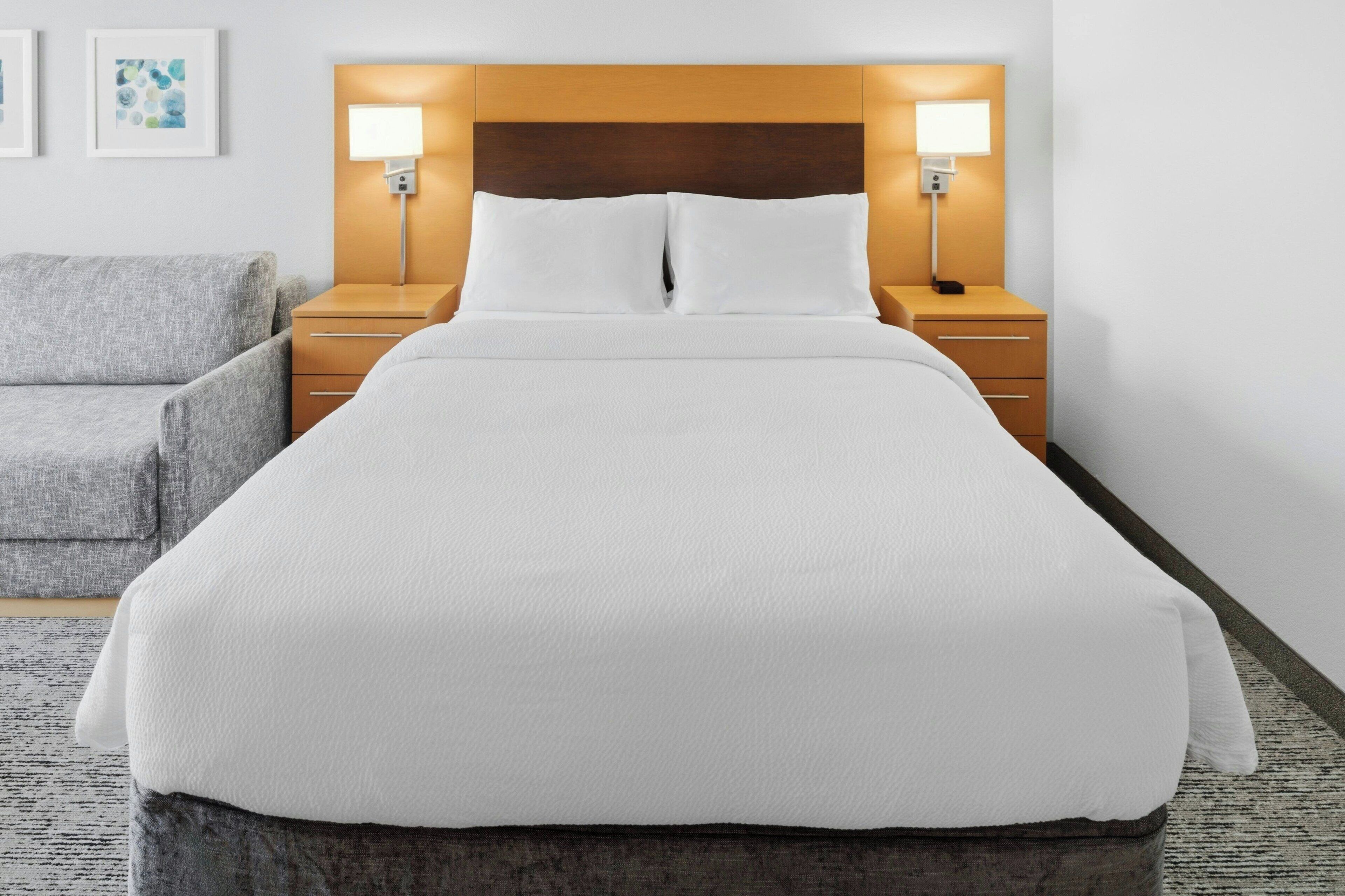 A neatly made bed with white linens, a wooden headboard, bedside lamps, and a gray armchair.