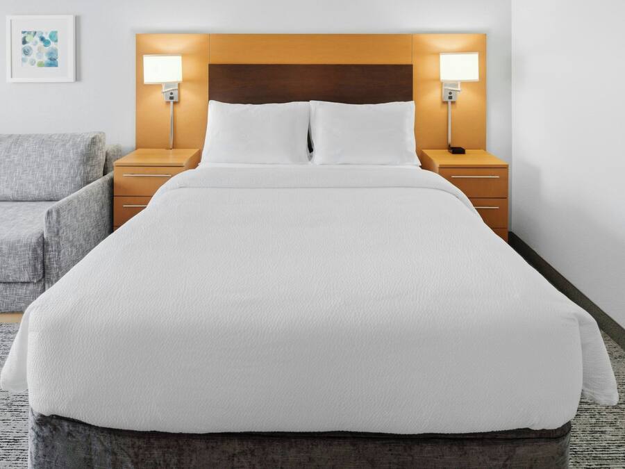A neatly made bed with white linens, a wooden headboard, bedside lamps, and a gray armchair.