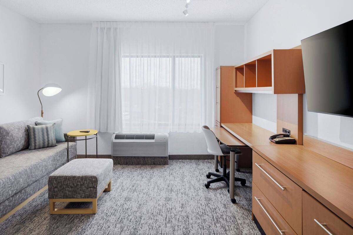 A modern hotel room with a sofa, ottoman, desk, chair, TV, and built-in shelving.