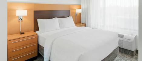 Una habitación de hotel con una cama grande, dos mesitas de noche con lámparas, un aire acondicionado instalado en la pared y una ventana con cortinas traslúcidas.