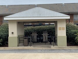 Un espacio al aire libre cubierto con una estación de parrilla, zona de estar y un edificio al fondo.