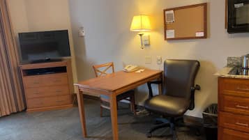 Habitación de hotel con escritorio de madera, una silla, televisor, lámpara, teléfono, un tablón de anuncios y una kitchenette.