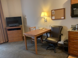 Habitación de hotel con un escritorio de madera, una silla, un televisor, una lámpara, un teléfono, un tablón de anuncios y una kitchenette.