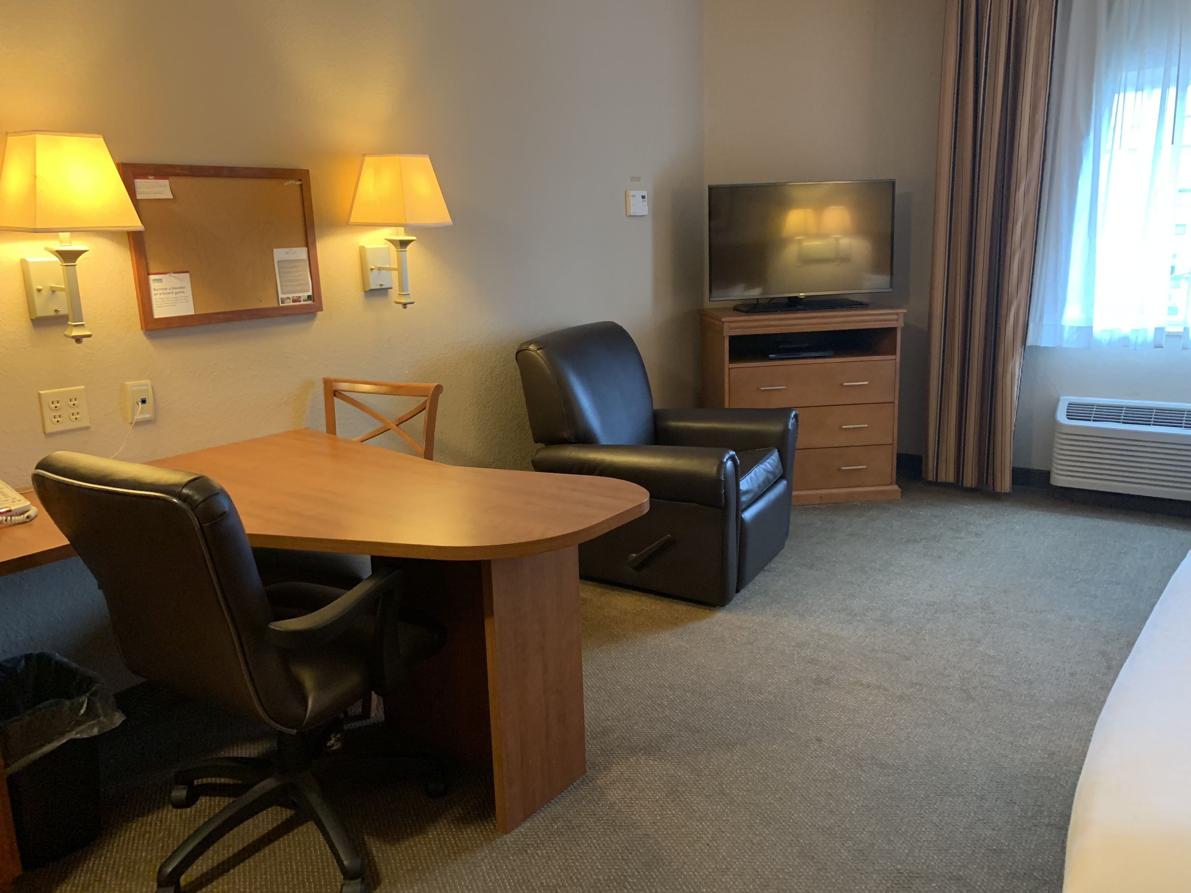 A hotel room with a wooden desk, two chairs, a television on a wooden cabinet, and a large window with curtains.