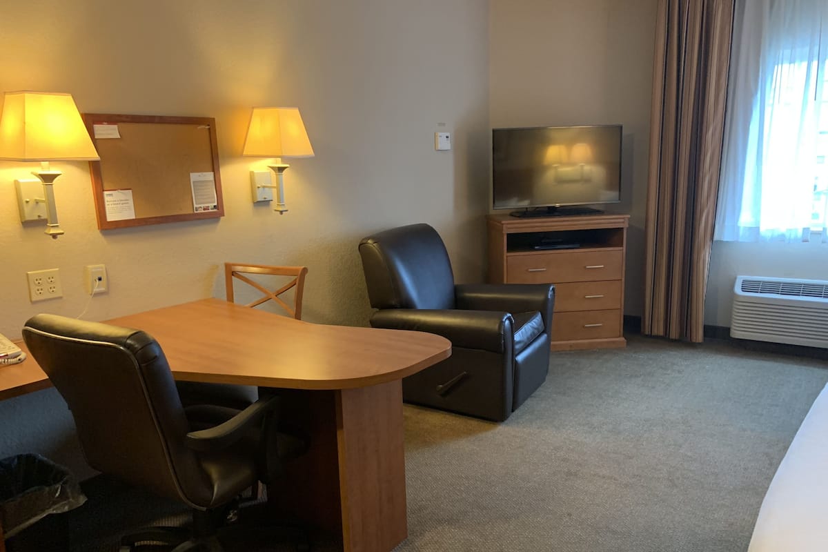 Habitación de hotel con escritorio de madera, dos sillas, un televisor sobre un mueble de madera y un ventanal con cortinas.
