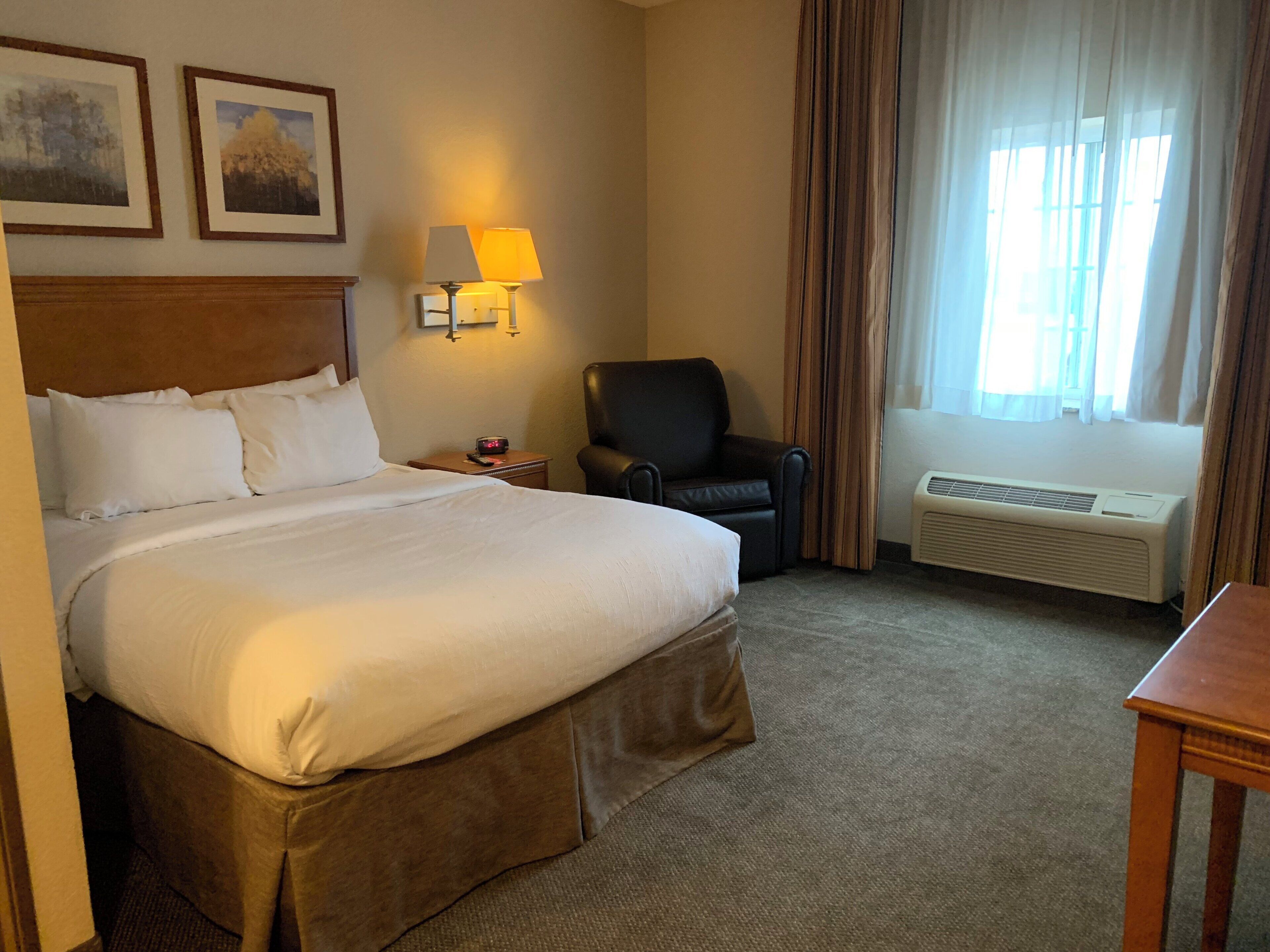 A hotel room with a large bed, a chair, a desk, a lamp, and a window with curtains.