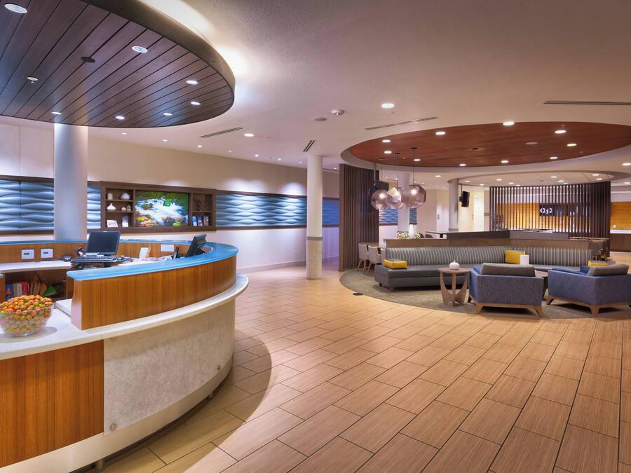 A modern hotel lobby with a reception desk, seating area, and a large screen displaying a scenic image.