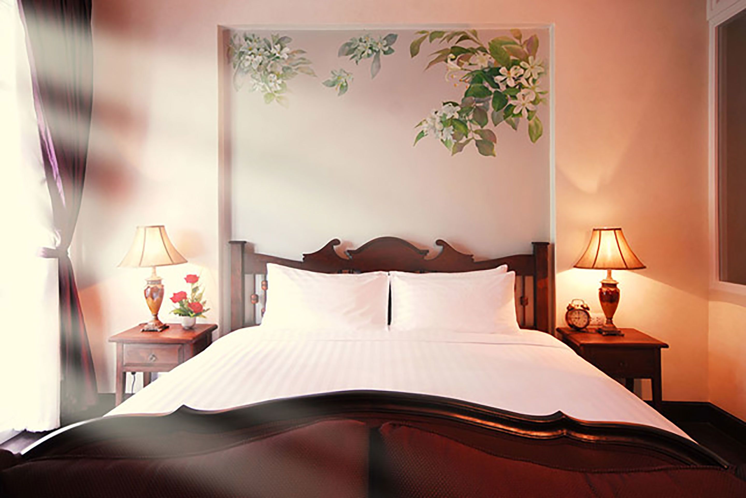 Una cama bien hecha con sábanas blancas, dos mesitas de noche con lámparas y un adorno floral en la pared.
