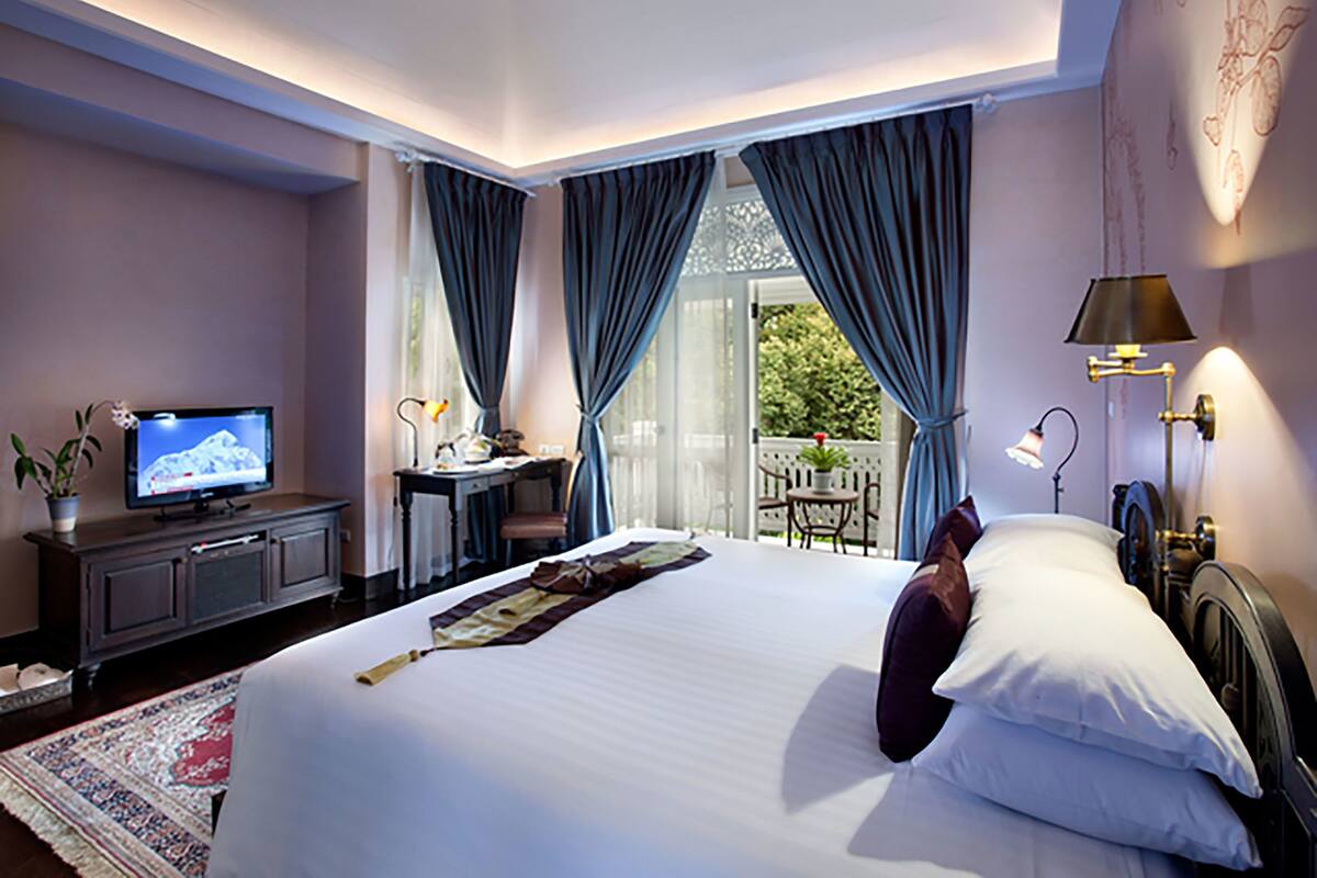 Une chambre d’hôtel avec un grand lit, un bureau avec un ordinateur, une télévision et un balcon donnant sur un paysage verdoyant.