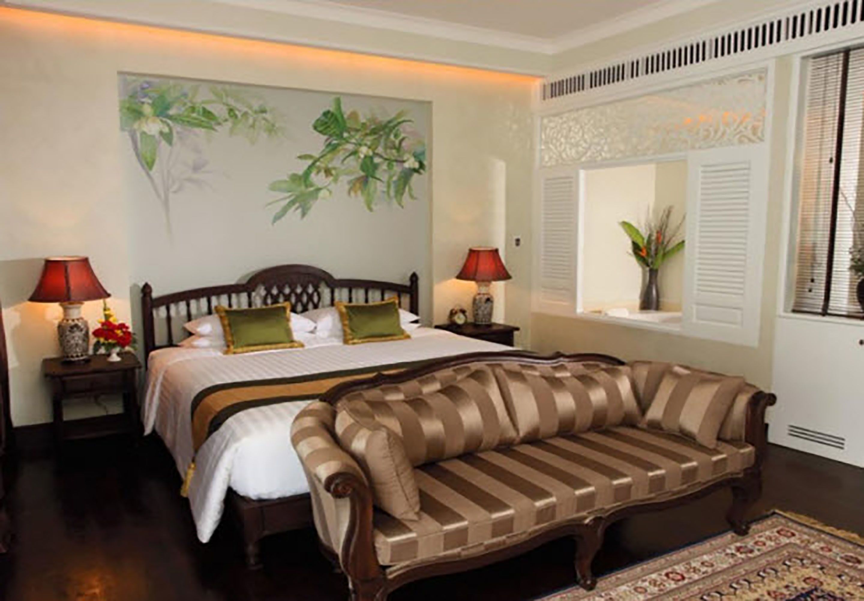 A bedroom with a bed, two bedside tables with lamps, a striped sofa, a window with shutters, and a painting on the wall.