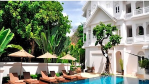 A pool area with lounge chairs and umbrellas, surrounded by lush greenery and a large white building with balconies.