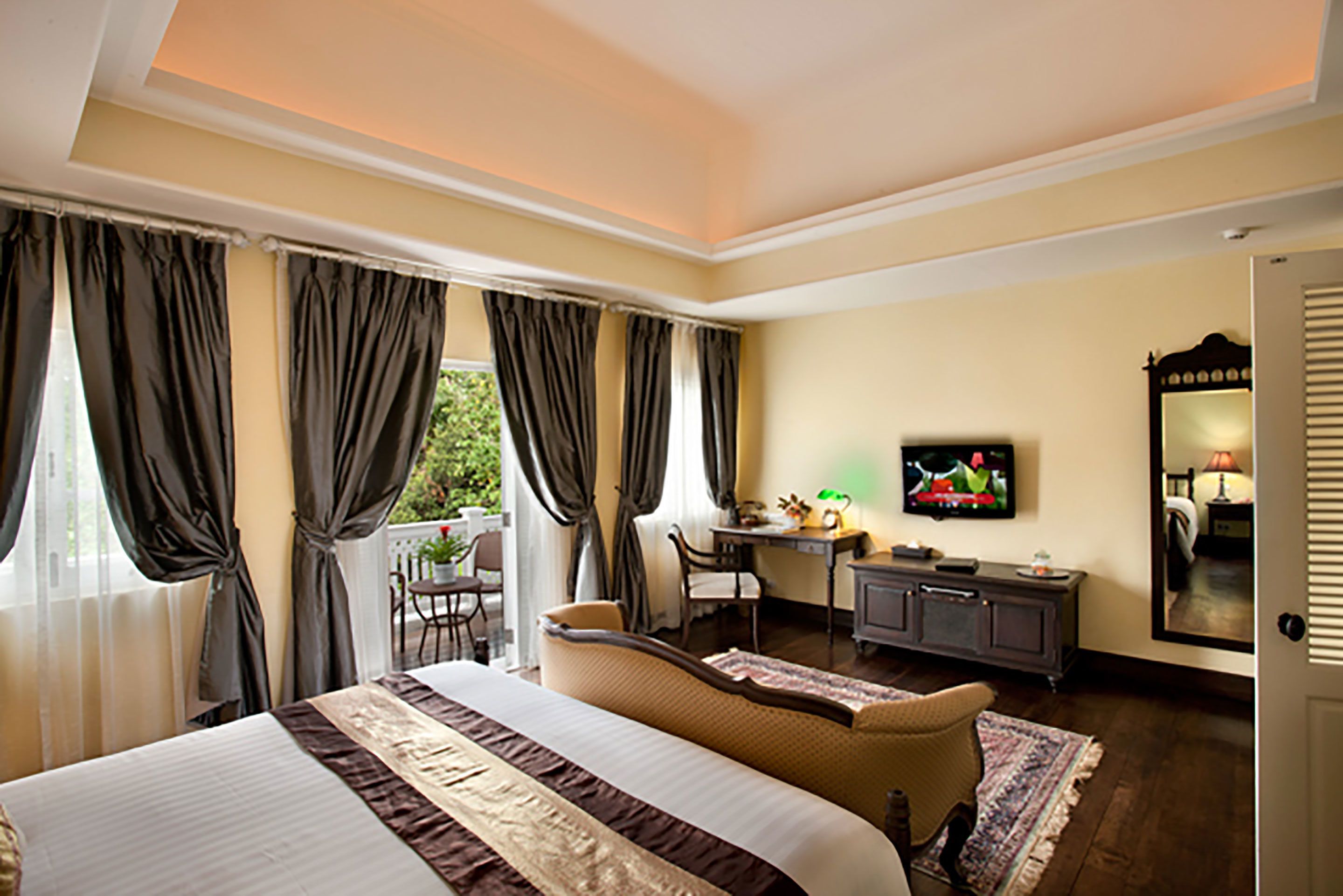 A hotel room with a large bed, a desk with a chair, a television, and a balcony with curtains.