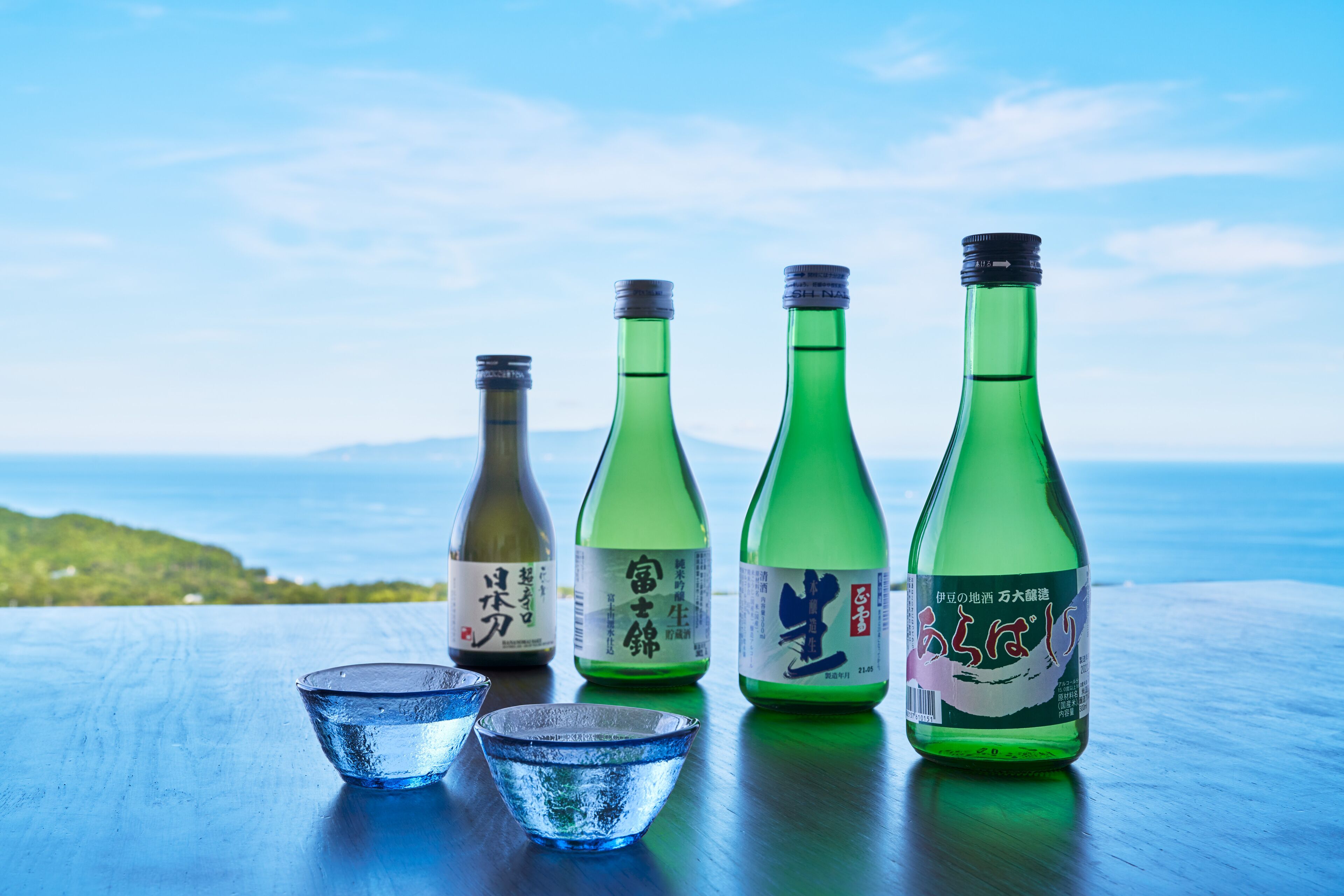 Vier groene flessen met etiketten, twee kleine blauwe schaaltjes, en een houten tafel met uitzicht op zee en heuvels op de achtergrond.