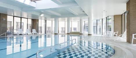Une piscine intérieure avec des parois en verre transparent, un sol à carreaux et des chaises blanches disposées autour de la piscine.