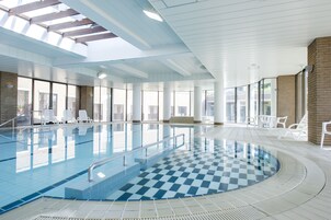 An indoor swimming pool with clear glass walls, a checkered floor, and white chairs around the pool.