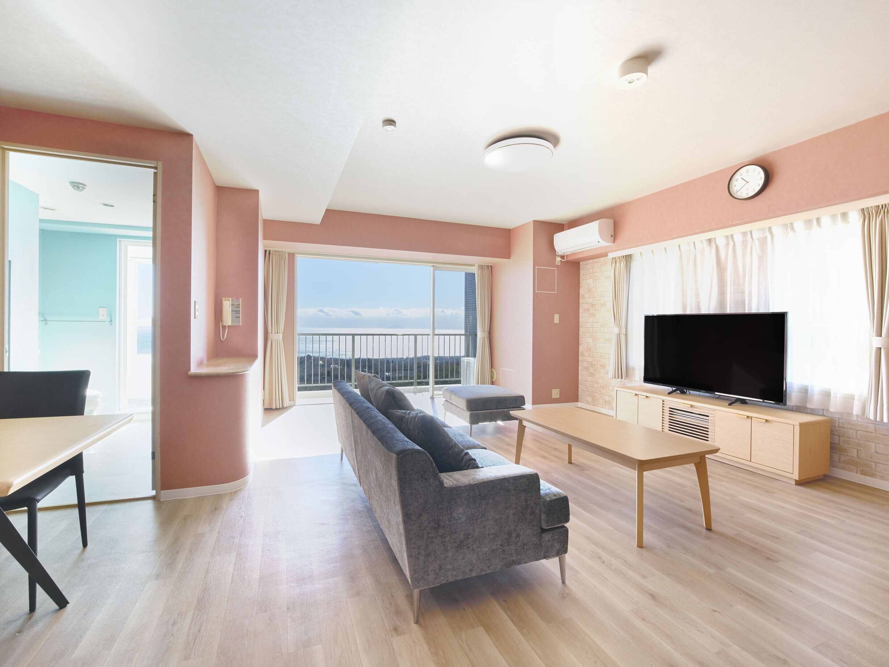 Una sala moderna con sofá, mesa de comedor y televisor. Hay un balcón con vista al mar.