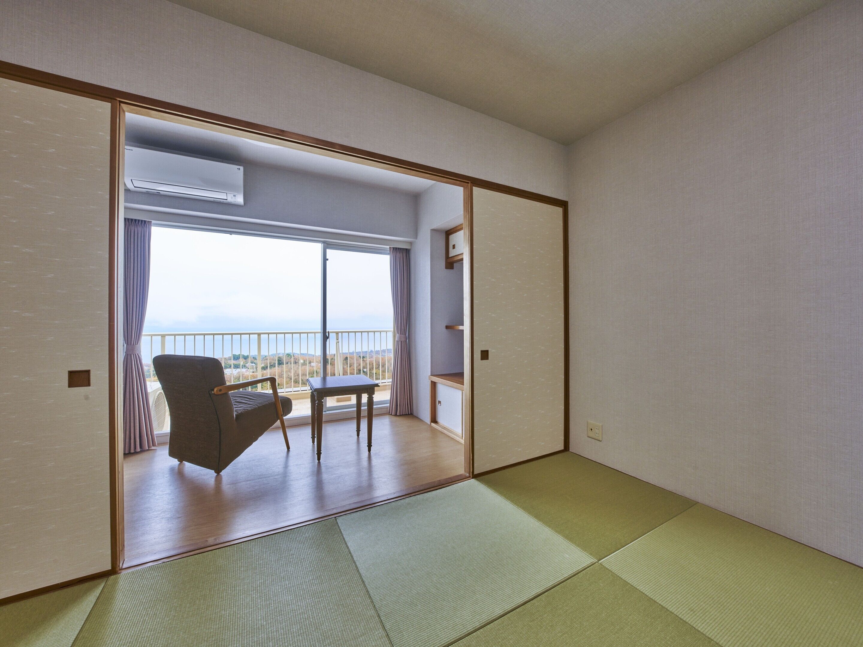 A room with tatami flooring, a sliding door leading to a balcony with a view, and a chair.