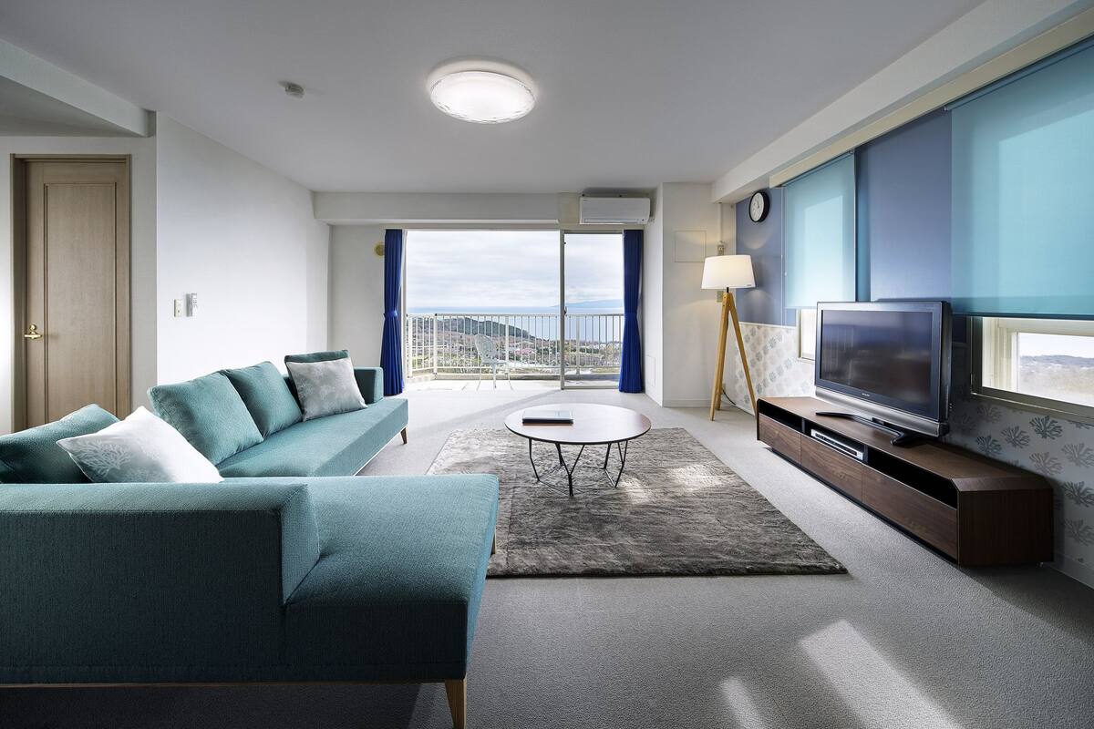 Un salon moderne avec un canapé, un téléviseur, une lampe et une vue sur la mer depuis un balcon.