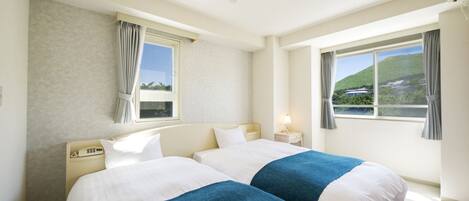 Ein Hotelzimmer mit zwei Betten, einem Nachttischtelefon, einem Beistelltisch mit Lampe und einem Fenster mit Blick auf eine grüne Landschaft.