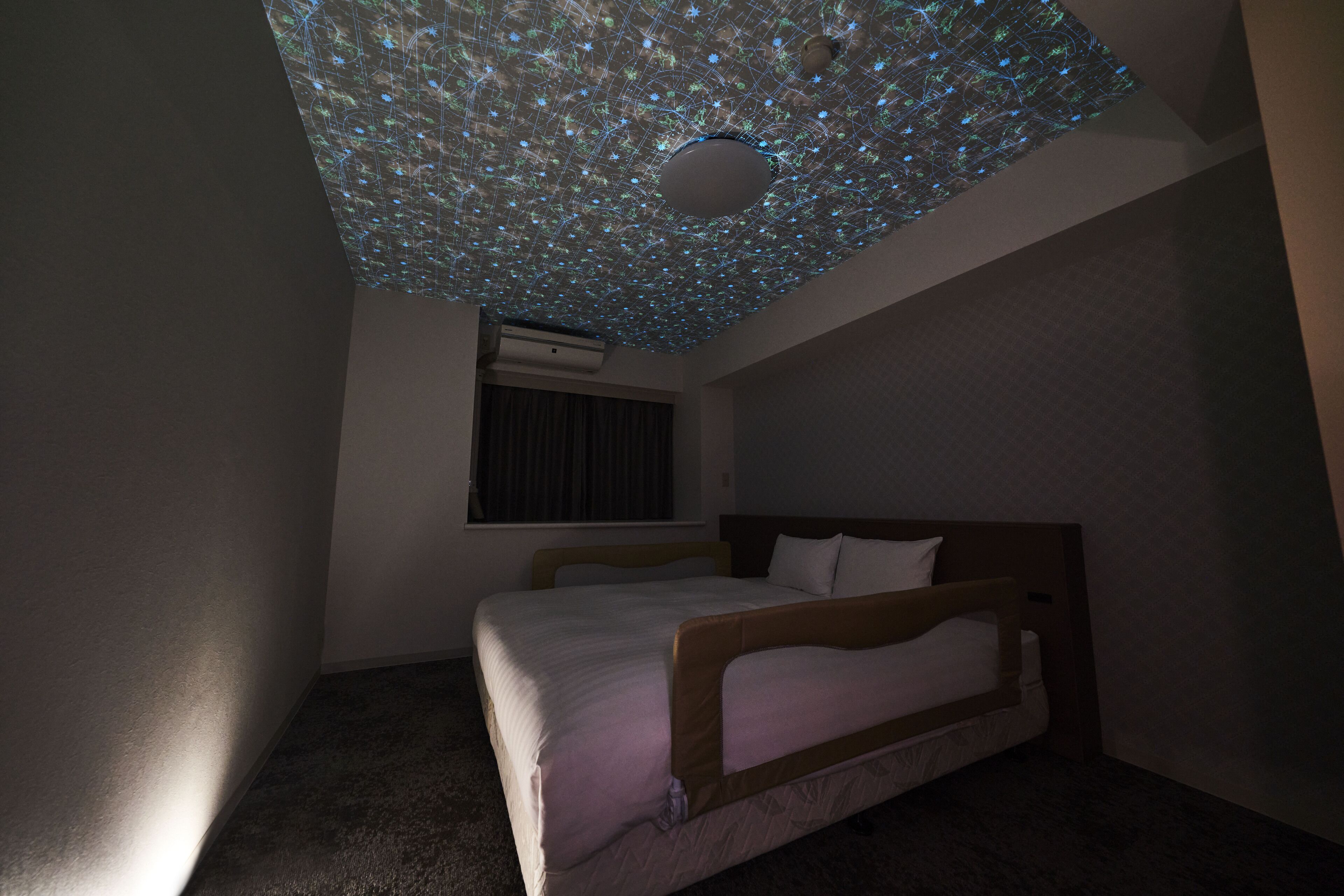 Un dormitorio con cama, ventana con cortinas y proyector de luz en el techo.