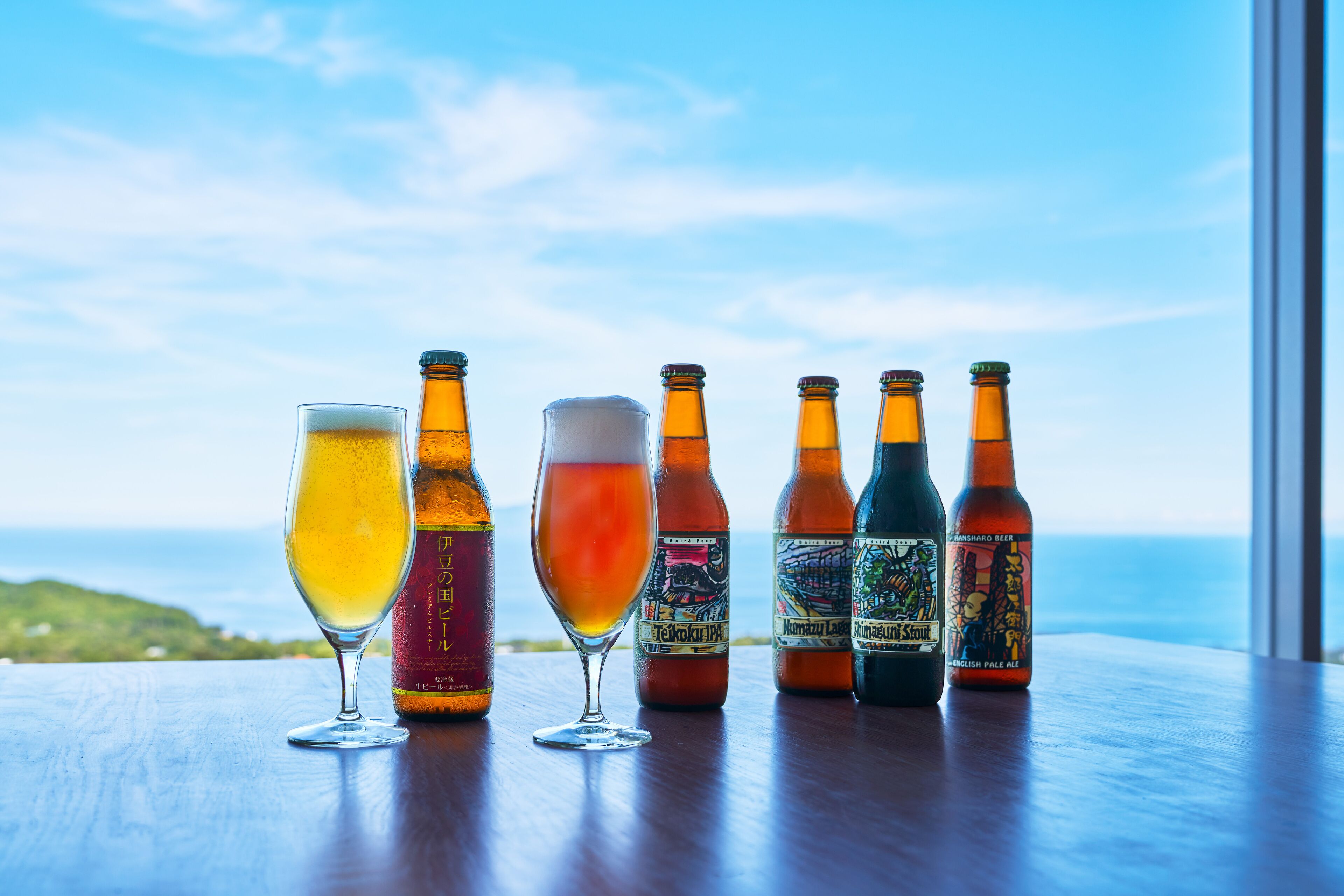 Een rij bierflesjes en glazen op een houten tafel, met uitzicht op de oceaan op de achtergrond.