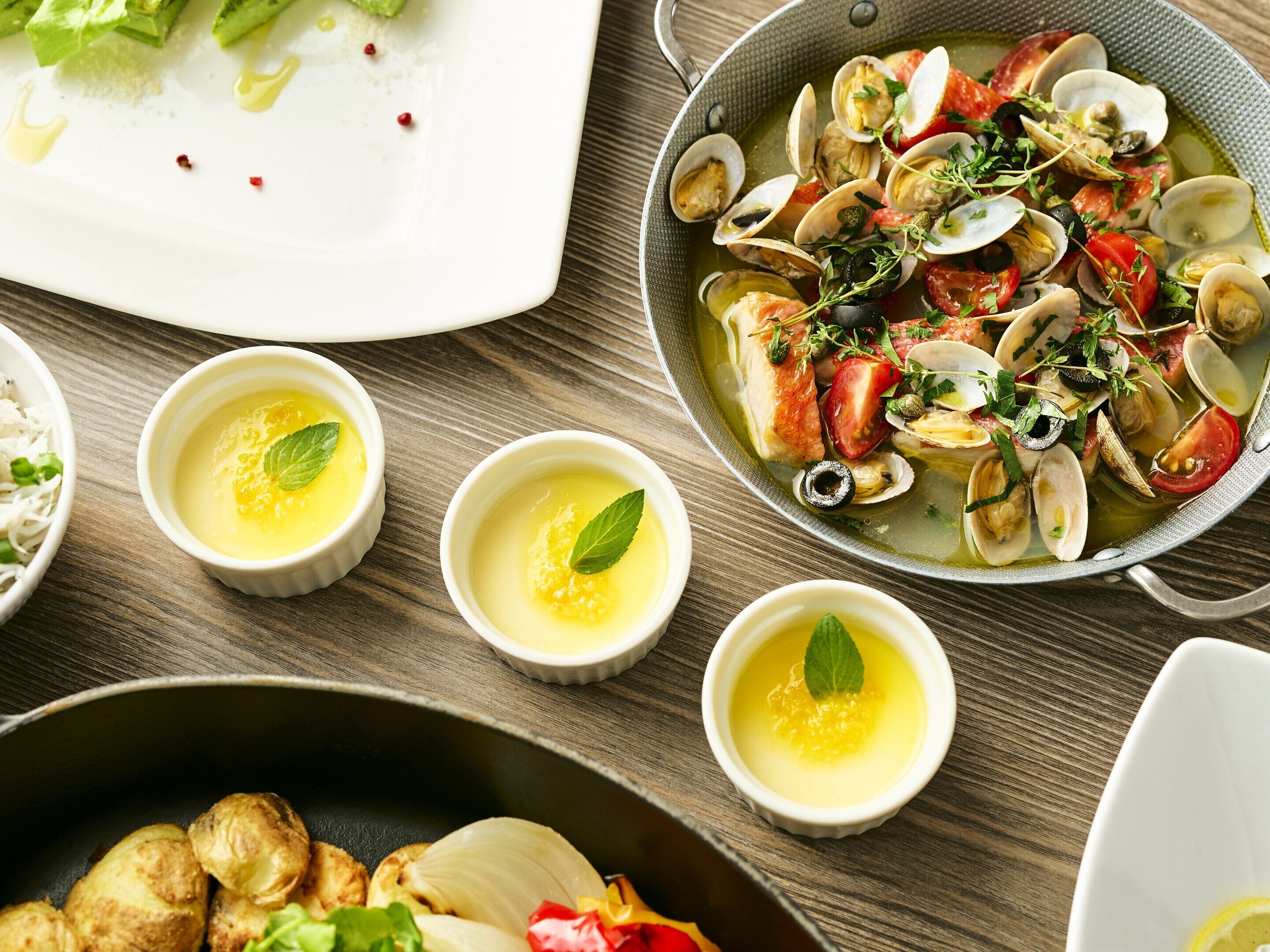 Un plato de mariscos con almejas y tomates, acompañado de pequeñas cazuelas de sopa y una fuente con verduras asadas.