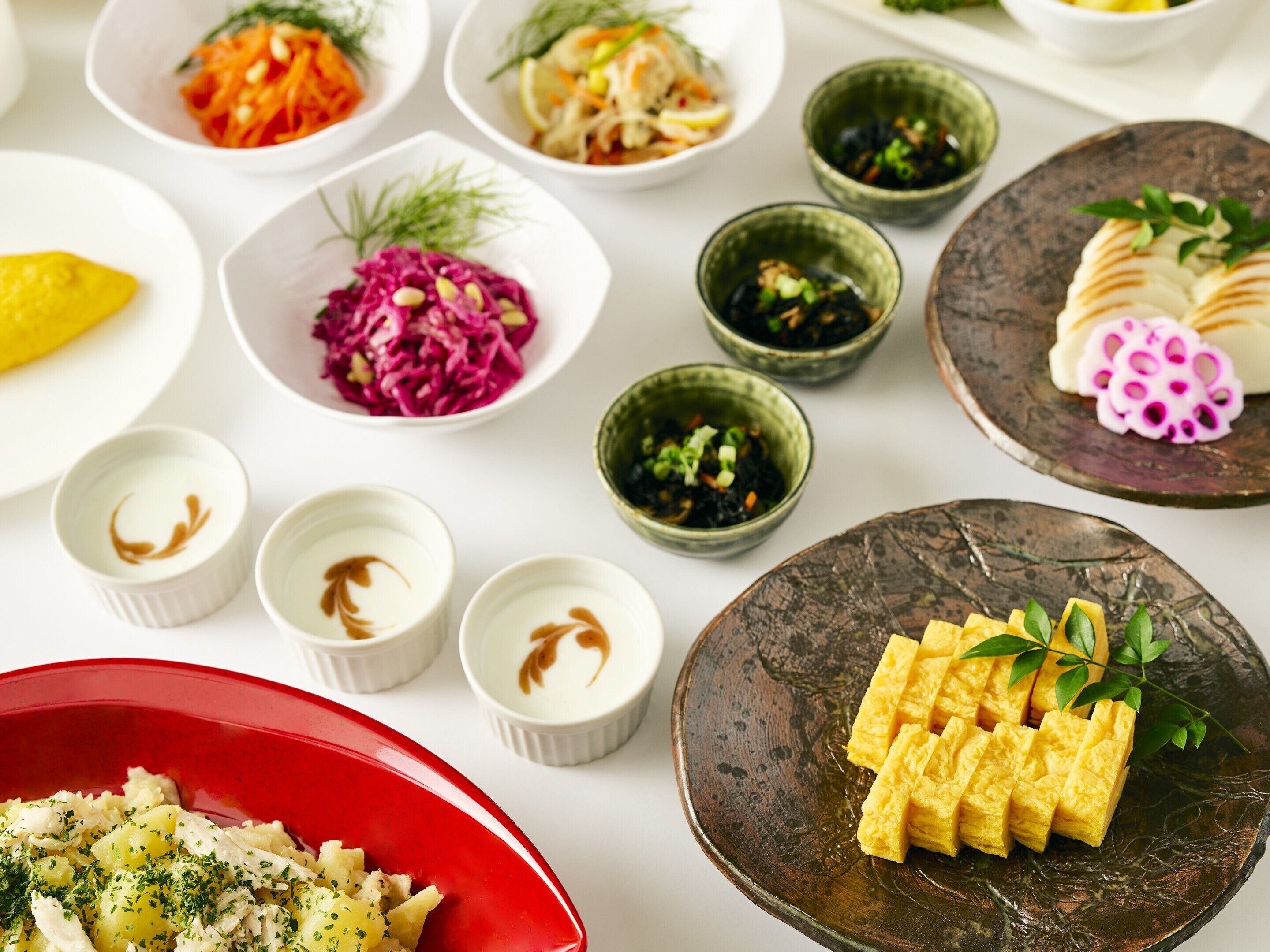 A variety of dishes including a red bowl of fish and potatoes, a plate of tofu, and several small bowls with different salads and sauces.